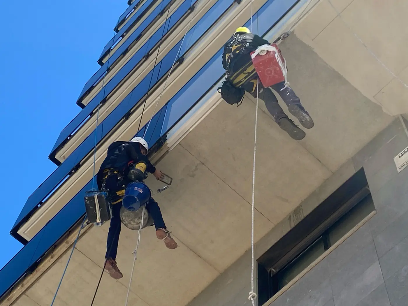 Dos técnicos colgados con cuerdas reparando los balcones de un edificio alto, sin andamio